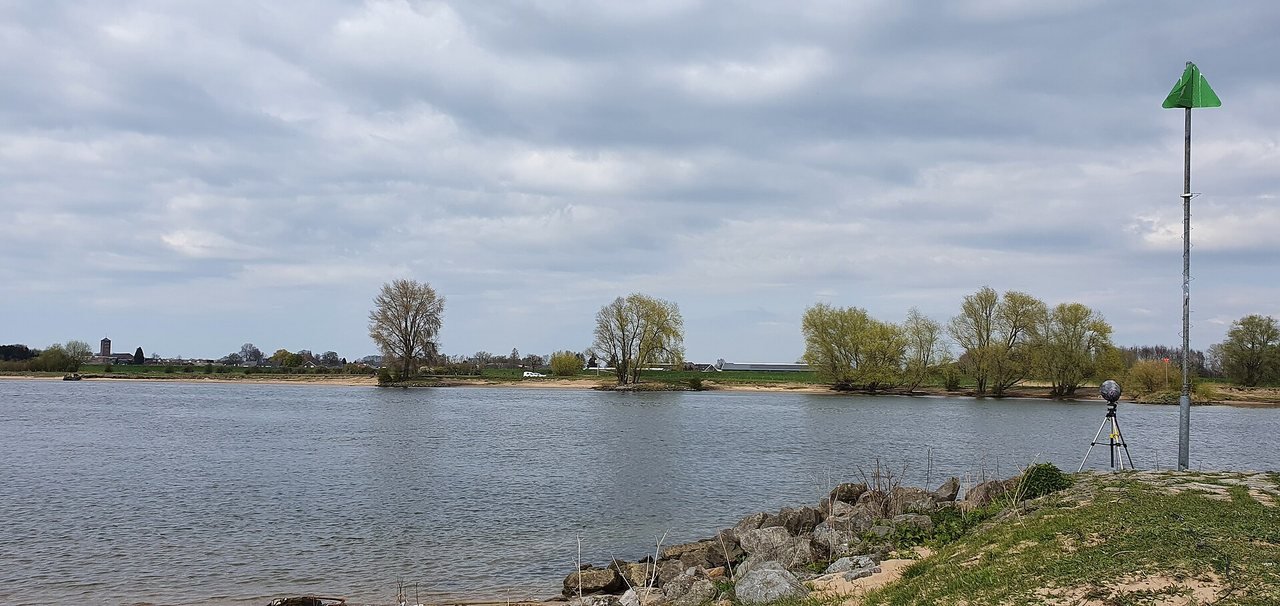 A wide, calm river under a grey-blue cloudy sky, with a low bank with willows on the far side and, in the foreground on the right, a grassy bank with stones, a green triangular post and a tripod.