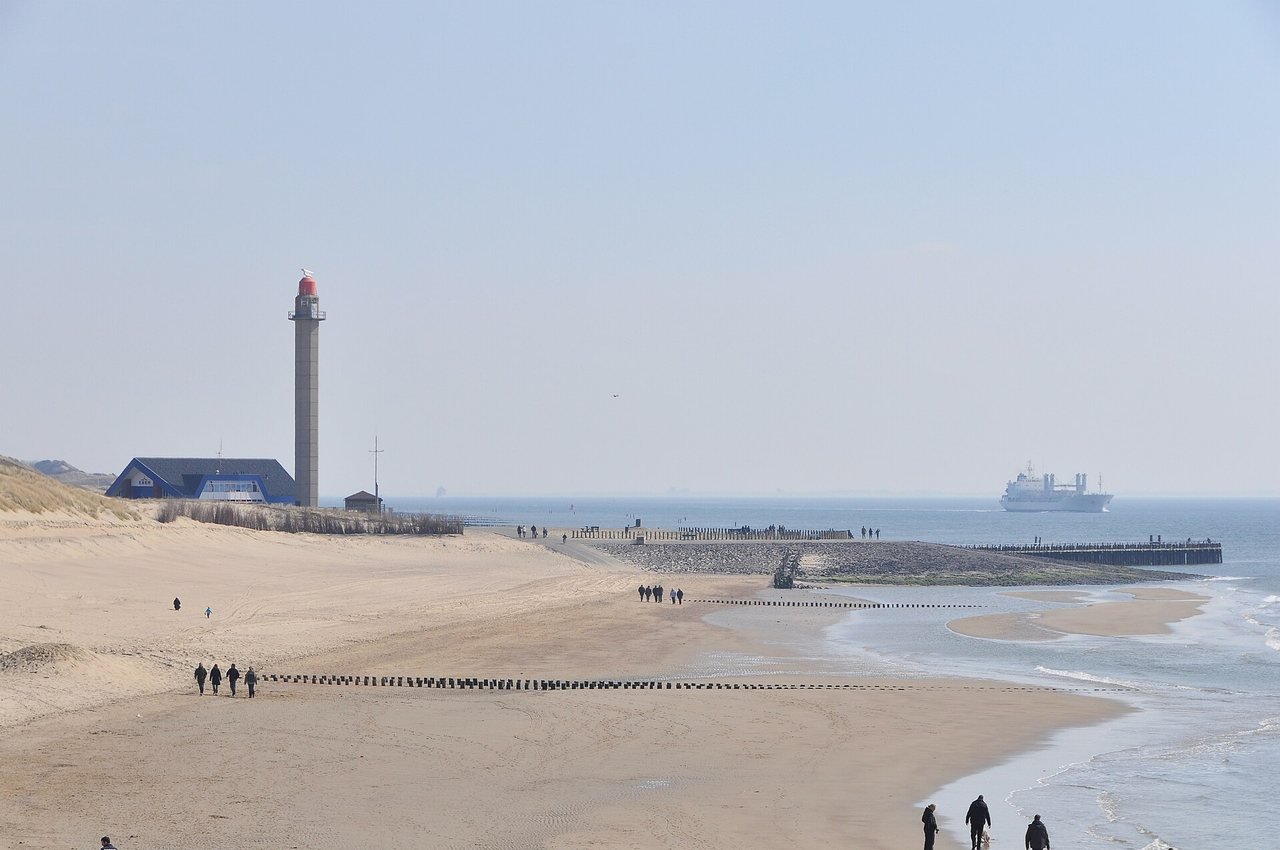 A wide sandy beach under a pale, hazy sky, with dunes, a blue building and a tall concrete lighthouse with a red top on the left, groynes and a stone breakwater in the middle, and a large white ship at sea in the background.