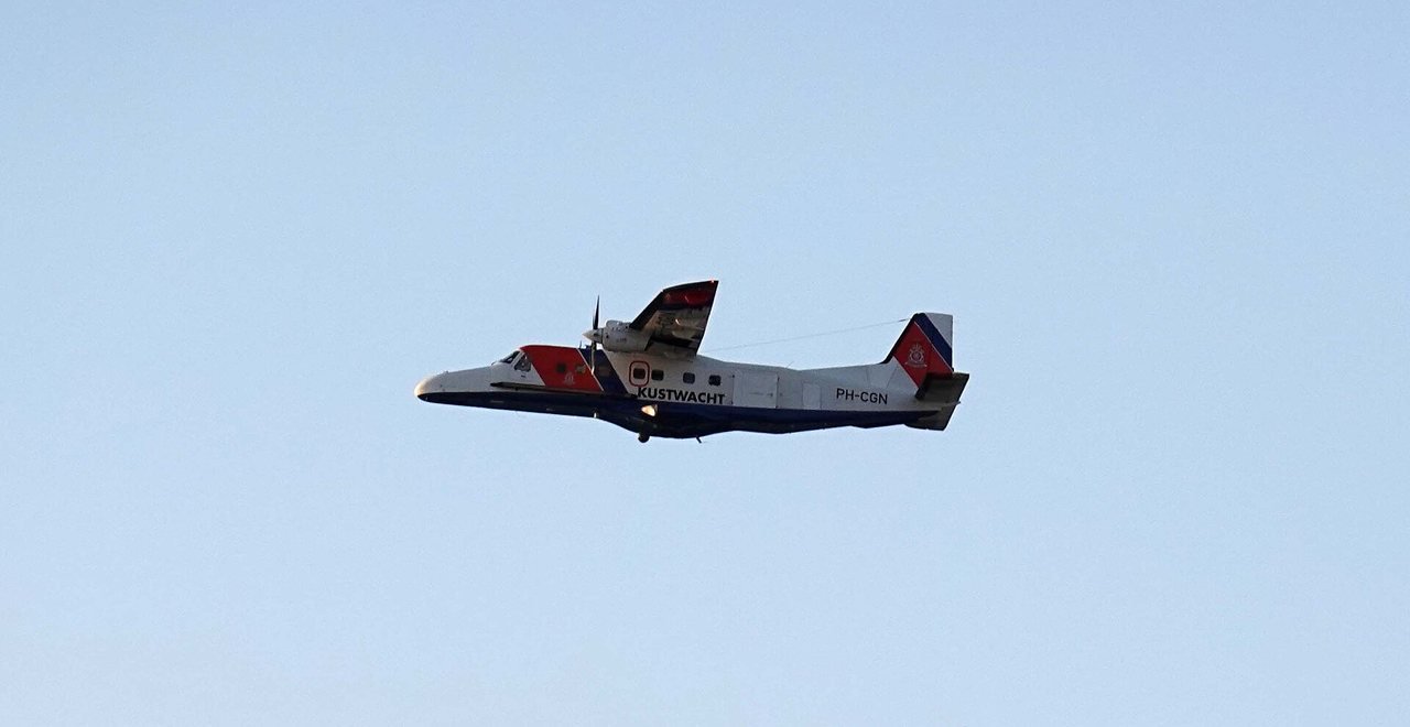 A white twin-engine propeller aircraft with red and blue stripes and the word Kustwacht (Coastguard) on the fuselage, in flight against an even pale blue sky.