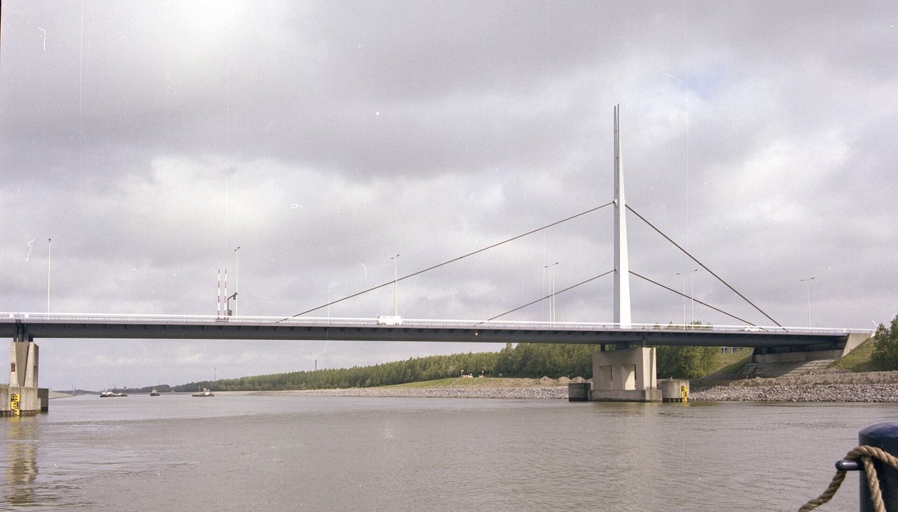 A long, low road bridge with a single white pylon and diagonal stay cables over a wide canal under a grey, cloudy sky, with a green bank with trees on the far side.