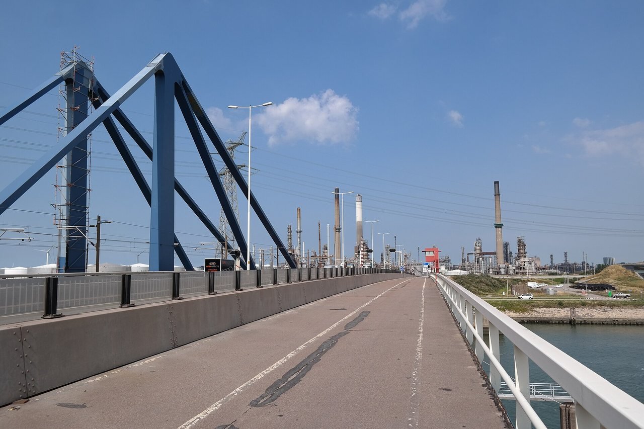 A wide cycle path over a bridge, with white railings on the right and a concrete barrier on the left, behind which stands a large blue steel pylon, under a blue sky with electricity pylons and factory chimneys on the horizon.
