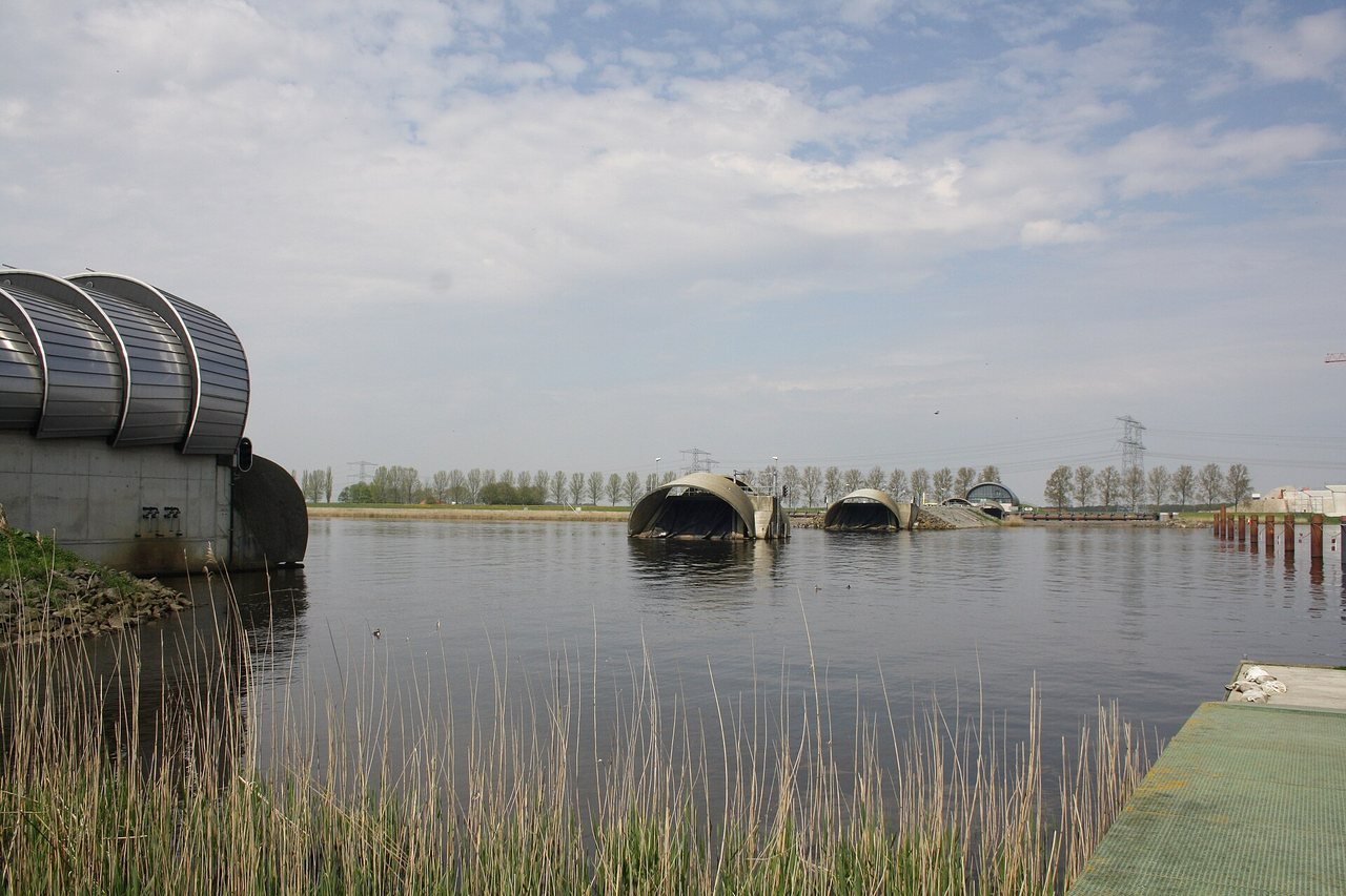 Wide water with reeds in the foreground; on the bank to the left a large building with curved metal roof segments, and further out in the water two smaller semicircular structures, under a light, cloudy sky.