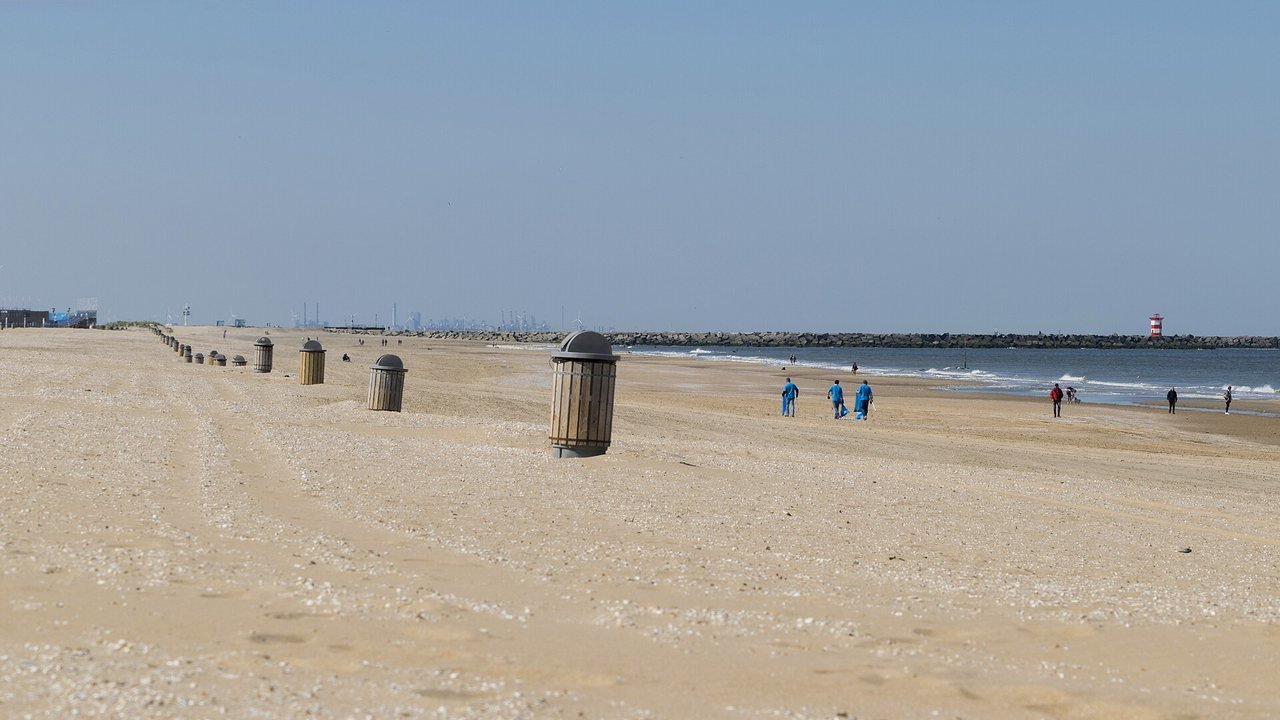 A wide, almost empty sandy beach under a pale blue sky, with a row of wooden litter bins on the left and, on the right, the sea, a stone harbour pier and a small red-and-white tower.