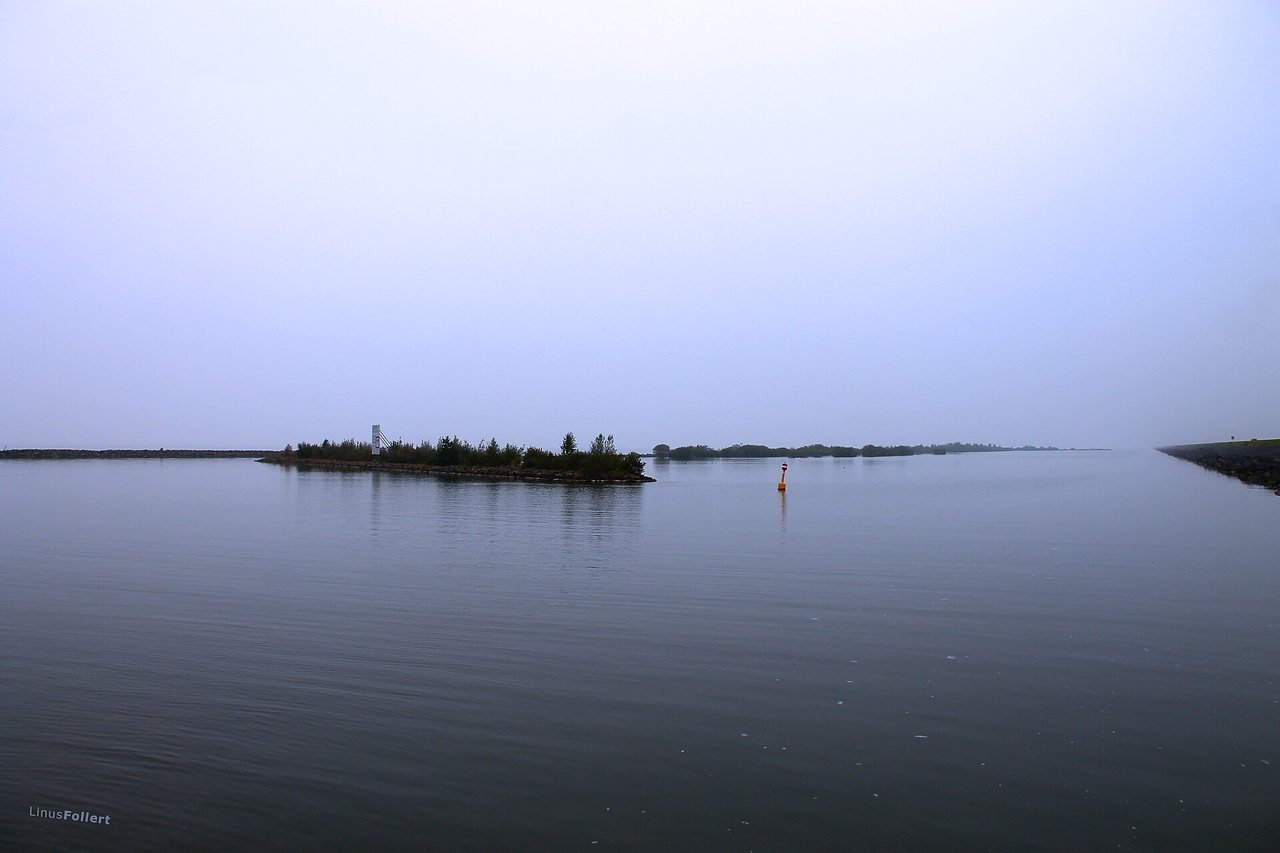 A mirror-smooth, grey-blue lake under an even, pale grey sky, with a low islet with trees in the middle, an orange buoy on the right and a narrow strip of land in the distance.