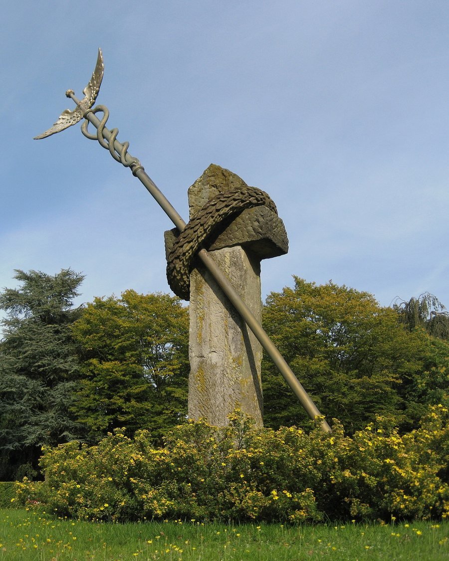 Hochformatfoto einer hohen Steinsäule mit einem gemeißelten Kranz, durch den ein langer, schräger Metallstab mit geflügelter Spitze ragt, vor Bäumen und gelb blühenden Sträuchern unter blauem Himmel.