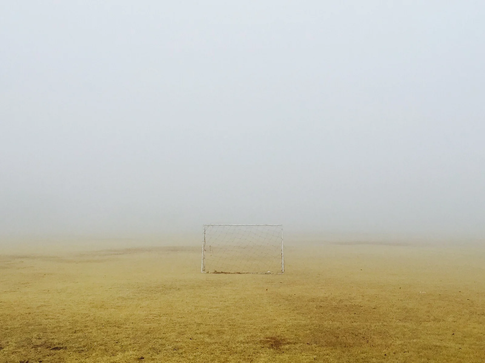Een wit doeltje met net midden op een geelgroen grasveld, omringd door dichte lichte mist.