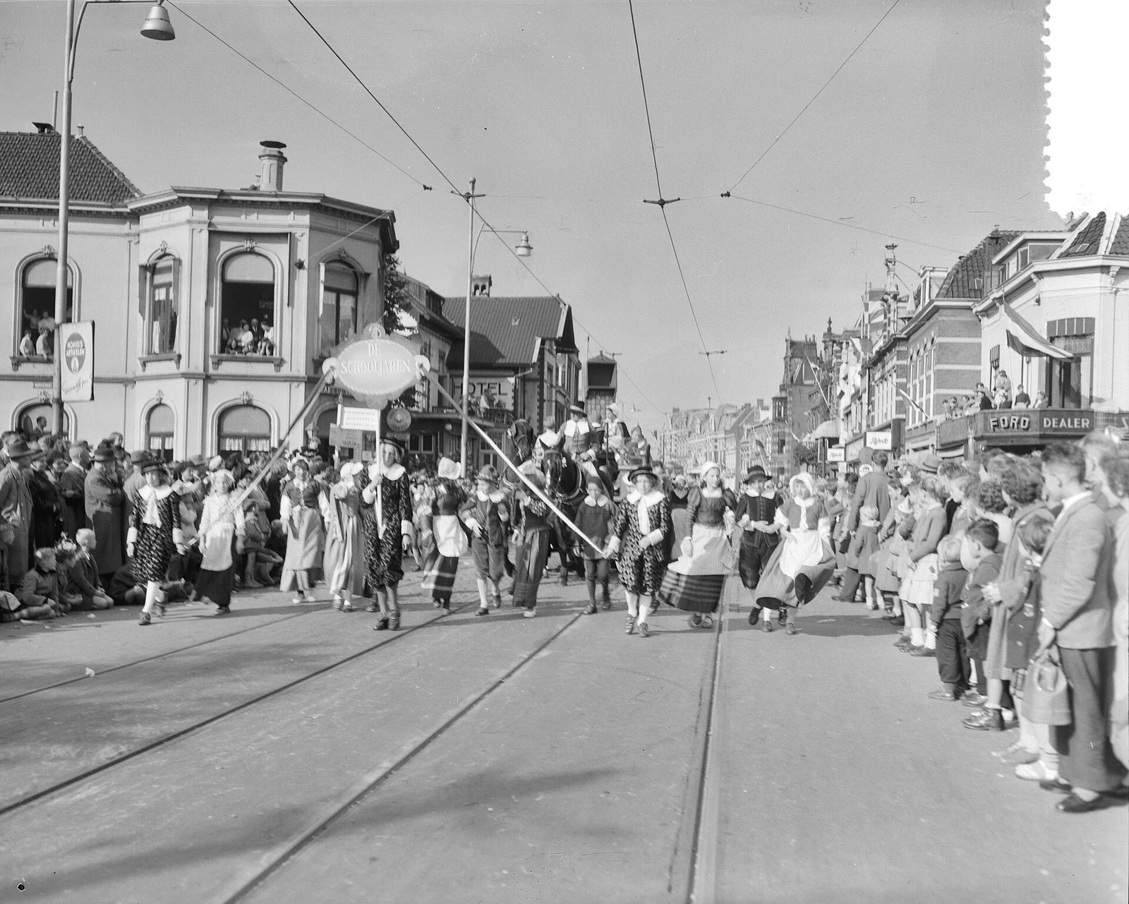 Zwart-witfoto van een optocht met mensen in kostuums en een ruiter, met aan weerszijden toeschouwers en huizen langs een brede straat met tramrails.