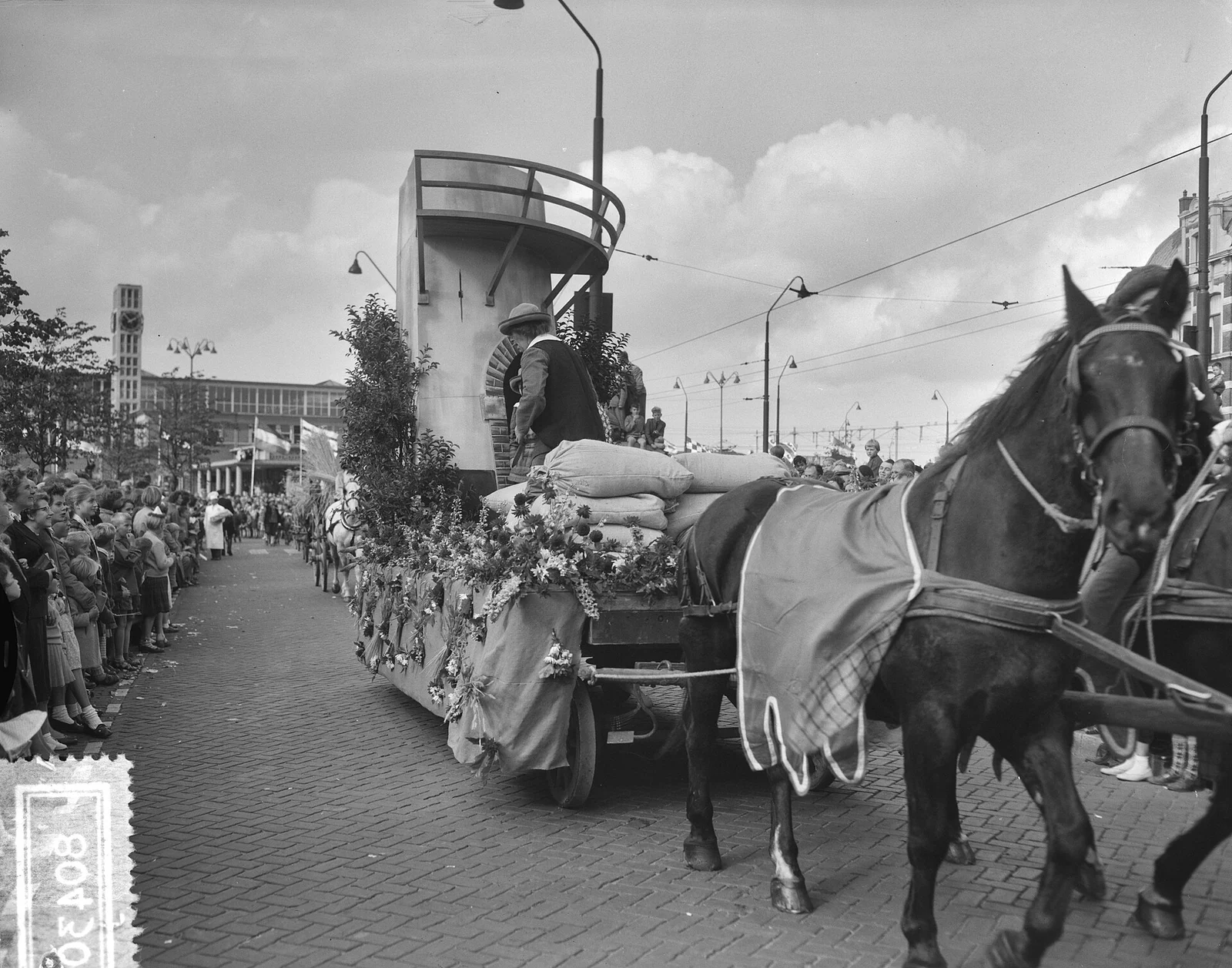 Zwart-witfoto van een met bloemen versierde wagen met zakken en een man in hoed, getrokken door paarden, met toeschouwers langs de straat.