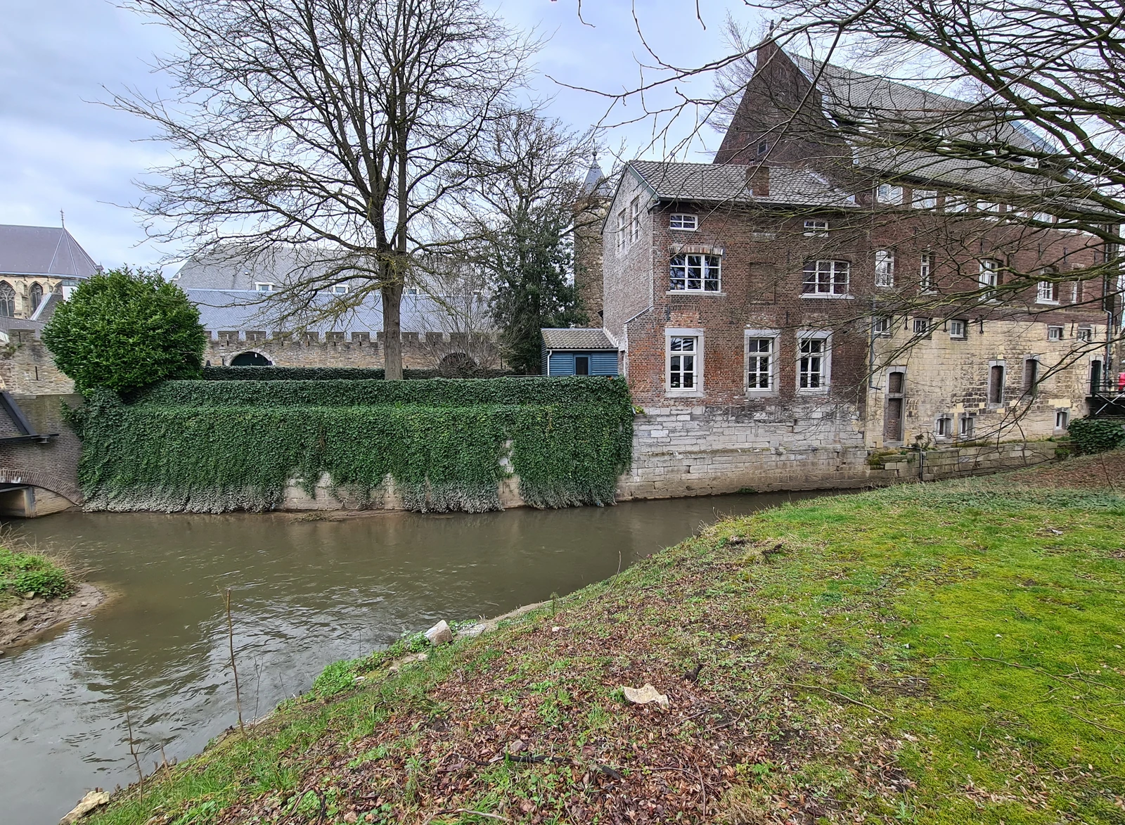 Een riviertje met bruin water stroomt langs een met klimop begroeide muur en een oud gebouw van baksteen en mergel, onder kale bomen.