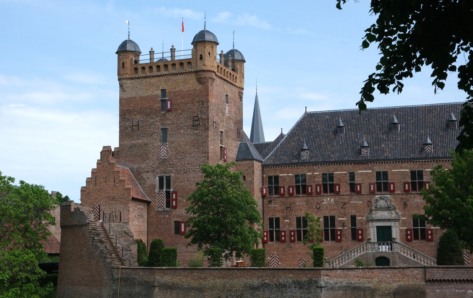 Een hoge vierkante bakstenen toren met kantelen en vier hoektorentjes, naast een vleugel met rode luiken en een stenen bordes, achter een bakstenen muur.